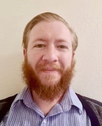 Therapist Cameron James standing indoors in front of a beige wall wearing a blue, striped button down shirt
