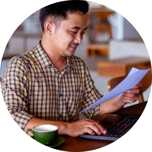 Man working with a laptop on a table. He is drinking coffee and looking at a piece of paper.
