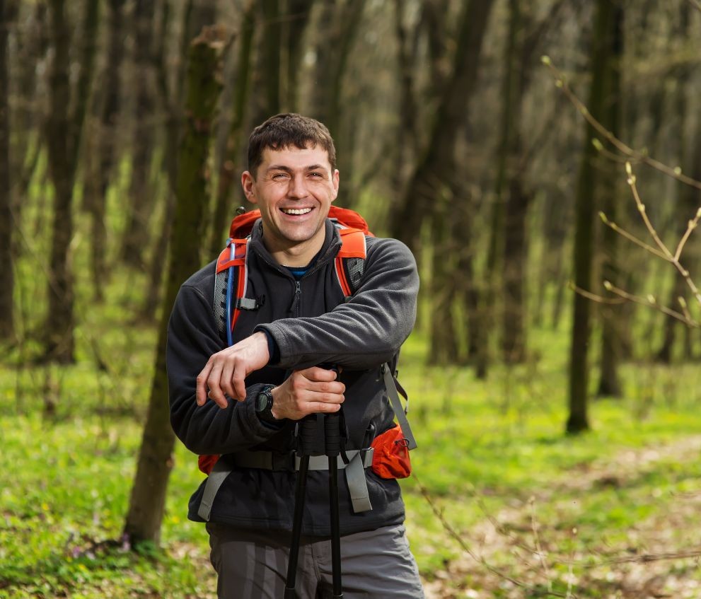 Happy man in hiking gear with a red backpack and trekking poles standing on a trail in a forest with fresh green foliage.